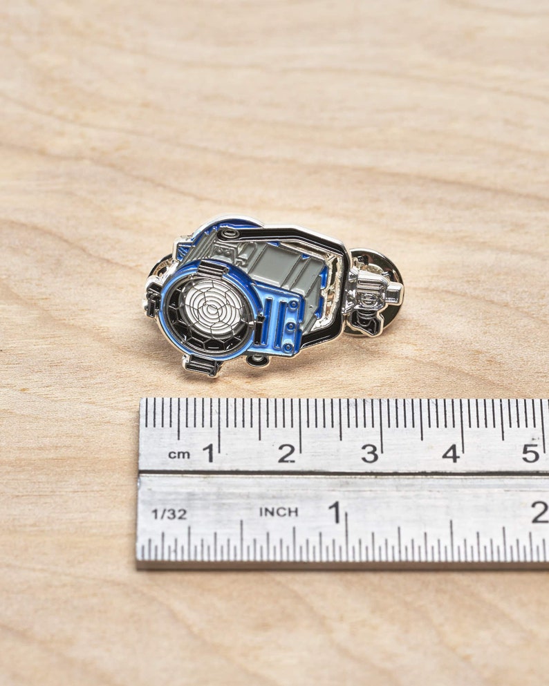 May include: A silver and blue enamel pin of a movie studio spotlight. The pin is about 1 inch wide and is sitting on a wooden surface next to a ruler.