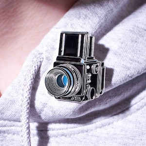 Puede incluir: Un pin de metal plateado con forma de cámara vintage. La cámara tiene una lente negra con un anillo azul alrededor.