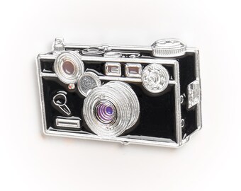 Puede incluir: Un pin de cámara de esmalte plateado y negro. La cámara tiene una lente plateada y un cuerpo negro. El pin es una excelente manera de mostrar tu amor por la fotografía.