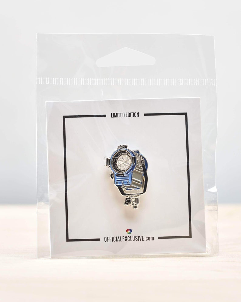 May include: A silver and blue enamel pin featuring a movie spotlight. The pin is packaged in a clear plastic bag with a white card that reads "Limited Edition" and "officialexclusive.com".