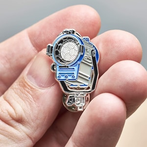 Puede incluir: Un pin de esmalte plateado y azul de un foco de cine. El pin tiene un diseño detallado con un mango negro y una luz blanca y azul.