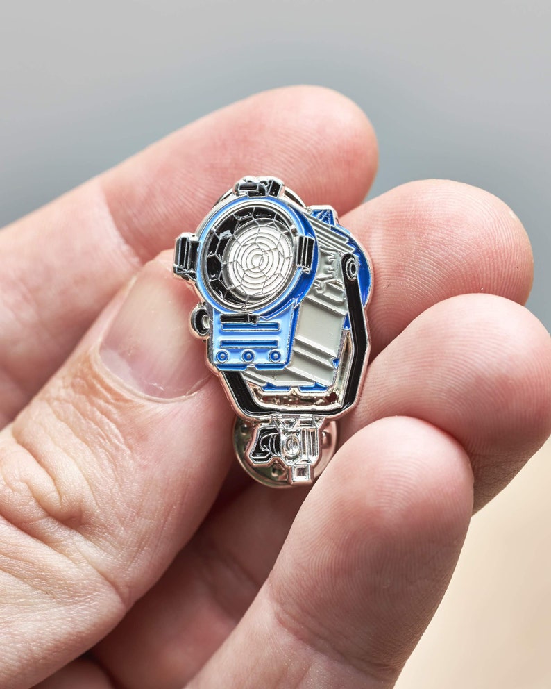 May include: A silver and blue enamel pin of a movie spotlight. The pin has a detailed design with a black handle and a white and blue light.