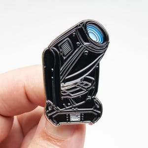 以下が含まれることがあります： ステージ照明器具の形をした黒いエナメルピン。光沢のある黒い仕上げに銀色の金属の輪郭、青と白の同心円状のレンズ、そしてベース部分には小さな長方形のスクリーンがデザインされています。