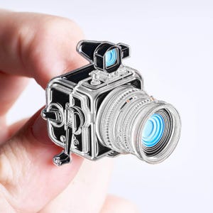 May include: A silver and black enamel pin shaped like a vintage medium format camera. The design features a prominent lens with blue circular accents, a top-mounted viewfinder with a blue interior, and a side-mounted winding crank handle.