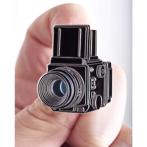 May include: A black enamel pin shaped like a vintage medium format camera. The design features a prominent lens with concentric circular detailing, a boxy camera body, and a top-mounted viewfinder. The metal outlines are silver-toned.