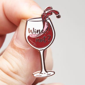 May include: A rose gold metal enamel pin shaped like a wine glass filled with deep red wine. The design features a splash of wine at the top and the text "Wine not?" written in black script across the centre of the glass against a white background.