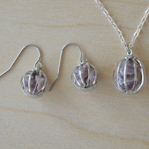 May include: A silver necklace and earrings set with a caged pink stone pendant and matching earrings.
