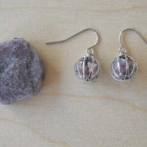 May include: A pair of silver earrings with a caged pink crystal stone. The earrings are hanging from a silver hook.