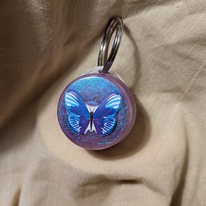 May include: A purple glitter keychain with a blue butterfly design. The keychain has a silver ring.