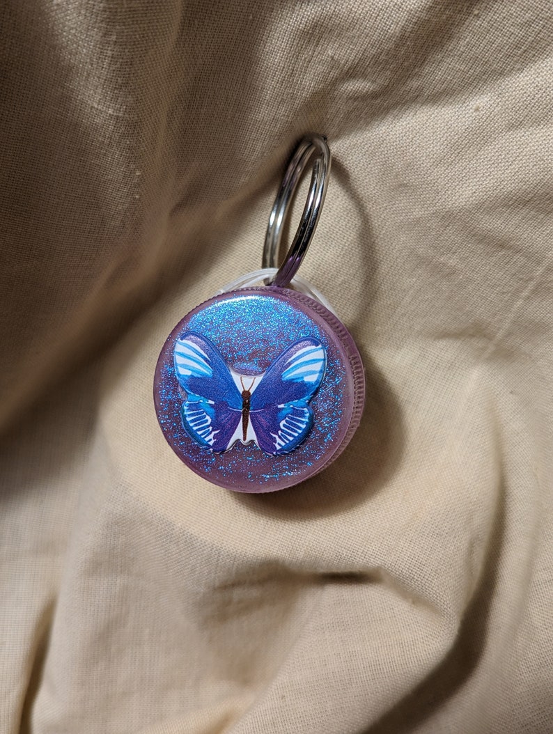 May include: A purple glitter keychain with a blue butterfly design. The keychain has a silver ring.