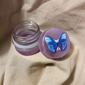 May include: A purple glass jar with a clear lid and a purple lid with a blue butterfly design.