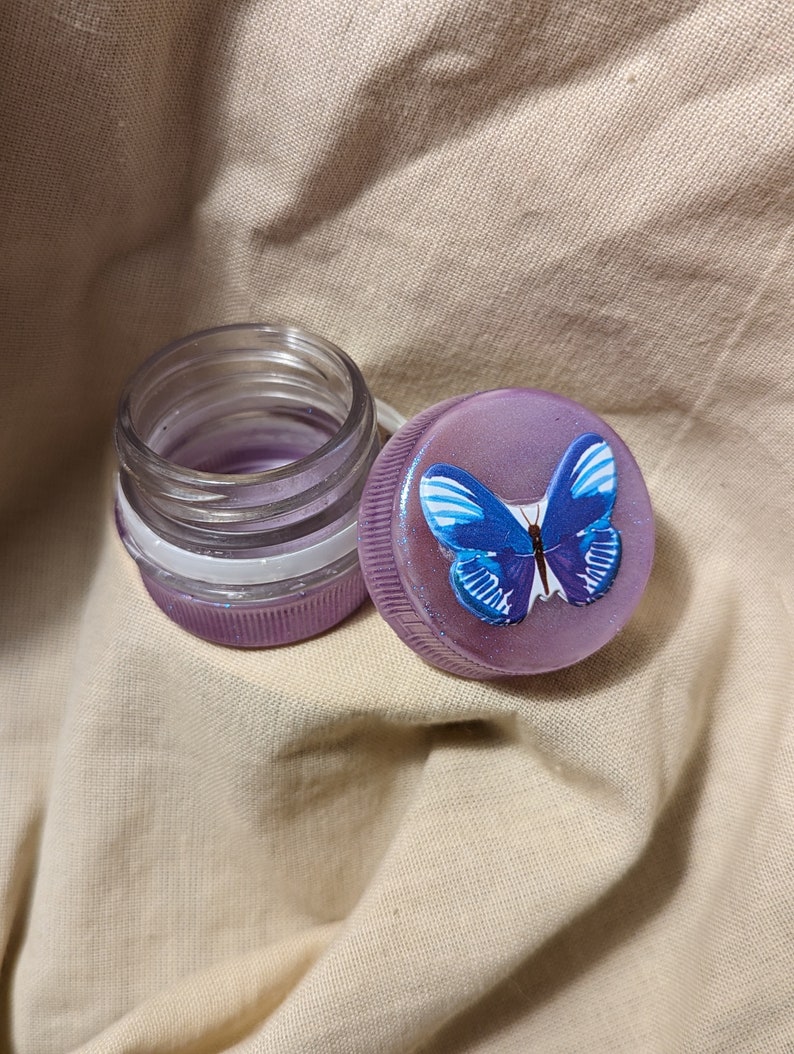 May include: A purple glass jar with a clear lid and a purple lid with a blue butterfly design.