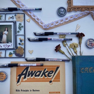 Peut inclure: Une image &agrave; plat d'une surface blanche avec divers objets, dont une photo encadr&eacute;e avec des fleurs s&eacute;ch&eacute;es, un livre intitul&eacute; "CREATION", un magazine intitul&eacute; "Awake!" avec le texte "Bible Principles in Business", quelques stylos et plusieurs petits boutons ronds avec des textes tels que "Best Life Ever" et "JW.org".