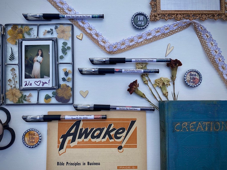 Peut inclure: Une image &agrave; plat d'une surface blanche avec divers objets, dont une photo encadr&eacute;e avec des fleurs s&eacute;ch&eacute;es, un livre intitul&eacute; "CREATION", un magazine intitul&eacute; "Awake!" avec le texte "Bible Principles in Business", quelques stylos et plusieurs petits boutons ronds avec des textes tels que "Best Life Ever" et "JW.org".