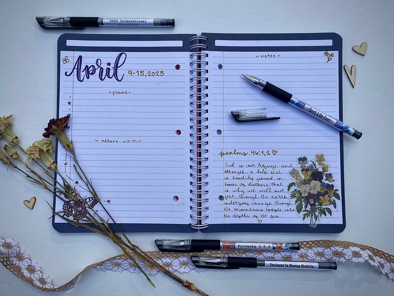 Peut inclure: Un agenda ouvert avec une couverture bleue et des pages blanches lign&eacute;es. L'agenda est ouvert sur avril 9-15, 2025. La page de gauche a le mot "avril" &eacute;crit en cursive violette et la page de droite a les mots "notes" et "psalmes 46:1,2" &eacute;crits en cursive noire. Il y a plusieurs stylos noirs et quelques fleurs s&eacute;ch&eacute;es sur la table.