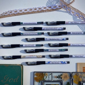 Peut inclure: Une collection de 10 stylos noirs avec des &eacute;tiquettes d&eacute;coratives comportant des citations inspirantes et des versets bibliques. Les stylos sont dispos&eacute;s en deux rang&eacute;es sur une surface blanche avec une dentelle et un livre en arri&egrave;re-plan.