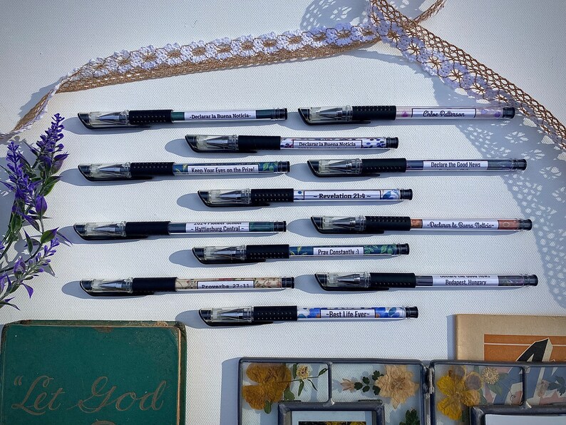 Peut inclure: Une collection de 10 stylos noirs avec des &eacute;tiquettes d&eacute;coratives comportant des citations inspirantes et des versets bibliques. Les stylos sont dispos&eacute;s en deux rang&eacute;es sur une surface blanche avec une dentelle et un livre en arri&egrave;re-plan.
