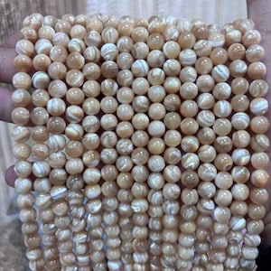 May include: Strands of round, beige mother-of-pearl beads. The beads have a pearlescent sheen with white and tan marbling. The beads are strung together, ready for jewelry making or other craft projects.