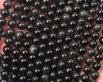 May include: A close-up of a collection of polished, round, black obsidian beads. The beads have a glossy finish and reflect light, creating highlights. The beads are strung together, suggesting they are for jewelry making or crafting.