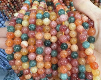 Puede incluir: Cadenas de cuentas de piedras preciosas de colores, en tonos naranja, rosa, verde y amarillo. Las cuentas redondas están ensartadas y se sostienen en una mano. El fondo muestra más cuentas de colores similares.