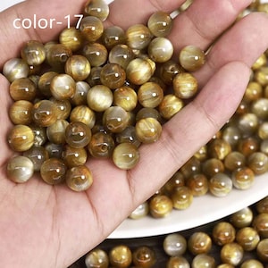 May include: A close-up of a hand holding a pile of golden sunstone beads. The beads are round and have a shimmery, iridescent finish. The beads are approximately 8mm in diameter.