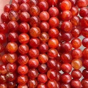 Könnte beinhalten: Nahaufnahme einer Sammlung roter und orangefarbener Edelsteinperlen. Die Perlen sind rund und poliert, mit einem durchscheinenden Aussehen. Die Farbe variiert von tiefem Rot bis zu hellem Orange, wobei einige Perlen eine Mischung aus beiden Farben aufweisen. Die Perlen sind aufgefädelt.