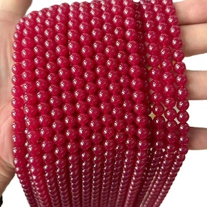 Puede incluir: Primer plano de hebras de cuentas redondas, brillantes, de color rojo intenso. Las cuentas son uniformes en tamaño y forma, y parecen estar hechas de un material liso y pulido. Las cuentas se sostienen en una mano, con un fondo borroso.