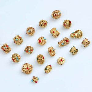 May include: Assortment of gold-toned metal beads with colorful gemstone accents. The beads feature intricate designs and various shapes, including round, cylindrical, and flower-shaped.