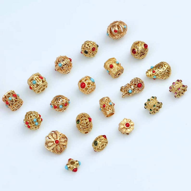 May include: Assortment of gold-toned metal beads with colorful gemstone accents. The beads feature intricate designs and various shapes, including round, cylindrical, and flower-shaped.