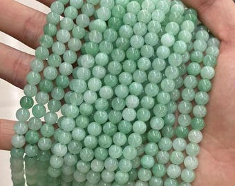 Puede incluir: Cadenas de cuentas redondas de color verde claro. Las cuentas son de tamaño y forma uniformes, con una apariencia ligeramente translúcida. Se sostienen en una mano, lo que sugiere que se exhiben o se ofrecen a la venta. La impresión general es la de una piedra preciosa o mineral natural.