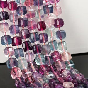 Könnte beinhalten: Eine Strähne quadratischer, irisierender Fluoritperlen in den Farben Lila, Rosa, Blau und Grün. Die Perlen sind auf einem durchsichtigen Faden aufgefädelt.