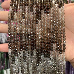 May include: Strands of faceted smoky quartz beads in varying shades of brown and clear. The beads are small and strung together, creating a shimmering effect. The beads are held in a hand, showcasing the color variations and the light reflecting off the cut surfaces.