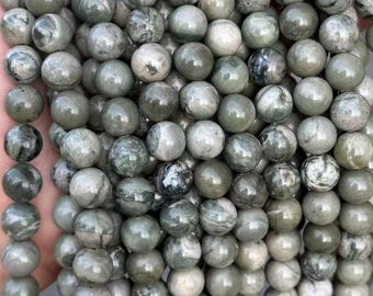 May include: Strands of round, polished beads in shades of gray and green. The beads have a marbled appearance, with variations in color and pattern. The beads are strung together, creating a textured surface. The beads are approximately 8mm in diameter.