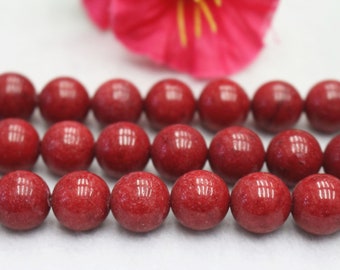 May include: Rows of round, deep red beads. The beads have a glossy finish and are arranged in neat rows. A blurred pink flower is visible in the background, adding a touch of color.