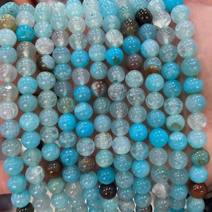May include: A collection of round gemstone beads in varying shades of blue. The beads range from pale to bright turquoise, with some displaying a crackled, translucent texture. Others are opaque. The beads are strung together, ready for crafting jewellery.