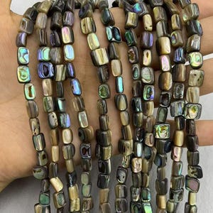May include: Strands of rectangular abalone shell beads in shades of brown, beige, and iridescent green and blue. The beads are strung together, creating a textured and colorful display, perfect for jewelry making.
