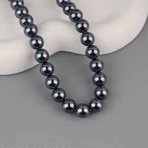 Op de afbeelding: Een ketting van donkergrijze, glanzende parels. De parels zijn rond en gelijkmatig van grootte, geregen tot een ketting. De ketting wordt getoond tegen een neutrale achtergrond, waardoor de kleur en glans van de parels worden benadrukt.