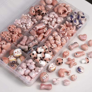 May include: A clear plastic storage box filled with various pink ceramic beads. The beads are shaped like animals, flowers, stars, and other designs. The box is open and some beads are scattered on the table.