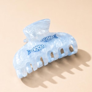 May include: A light blue hair claw clip with a marbled, translucent finish. The surface features two small, dark blue fish illustrations, each decorated with a delicate white floral pattern across their bodies. The clip has a curved, multi-pronged design.