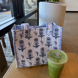 May include: A white tote bag with a blue floral pattern and blue striped trim sits on a wooden table next to a green matcha drink in a clear plastic cup. The bag has two handles.