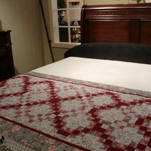 May include: A white bed with a red and white quilt on top. The bed is made of wood and has a headboard with intricate carvings. The bed is in a bedroom with a dresser and a window in the background.