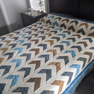 Op de afbeelding: Een wit, blauw en bruin chevron-patroon quilt met een gestikt ontwerp. De quilt ligt op een bed met een donkerbruine hoofdeinde.