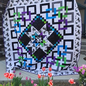 Puede incluir: Una colcha blanca con un diseño geométrico en blanco y negro en el centro. La colcha está rodeada por un borde de cuadrados blancos con caballos blancos y negros impresos en ellos. La colcha está colgada en una pared con una fila de tulipanes en primer plano.