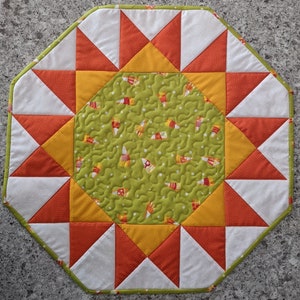 May include: Octagonal table runner with a green center featuring a repeating pattern of candy corn. The center is surrounded by a ring of alternating orange and yellow triangles with white triangles in between.