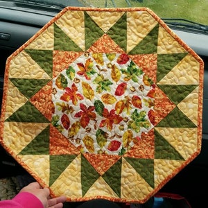 May include: Octagonal table runner with a central starburst design featuring a fall leaf print in shades of red, orange, yellow, and green. The outer border is a patchwork of yellow, orange, and green triangles.
