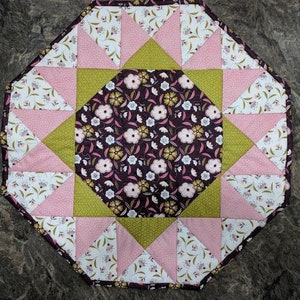 May include: Octagonal table topper with a starburst design. The centre is a brown fabric with white and green floral print. The starburst is made of pink and green fabric with white floral print.