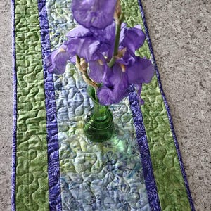 May include: A quilted table runner with a floral pattern in shades of green, blue, and purple. The runner has a green border and a purple center stripe. A small green vase holds purple iris flowers.