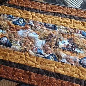May include: Quilted table runner with a rural landscape design, showing barns, trees, and horses. The colour scheme includes shades of brown, gold, and blue. The runner has a quilted texture with decorative stitching.
