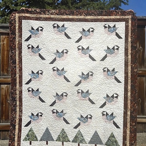 May include: A handmade quilt featuring a repeating pattern of stylized birds in blue, pink, and gray, with black wings and shadows. The quilt has a white background and a brown border with a floral design. At the bottom are green and gray trees.
