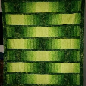 May include: A handmade quilt with a vibrant green color scheme. The quilt features horizontal bands of varying shades of green, from light lime to deep forest, creating a textured, layered effect. The edges are finished with a darker green border.